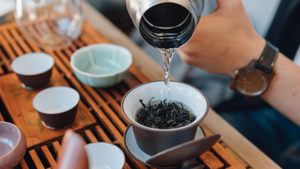 Person pouring tea from a silver teapot into a white bowl on a wooden tray.